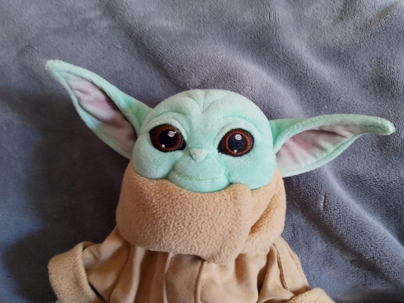 May include: A plush toy of a character with large ears, big brown eyes, and a light green face. The character is wearing a tan coat and a tan scarf. The toy is on a grey surface.