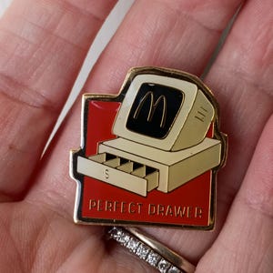 May include: A gold-toned pin featuring a computer with the McDonald's golden arches logo on the screen. The computer sits on a red background with the words "PERFECT DRAWER" in white. The drawer is open, revealing the letter "S".