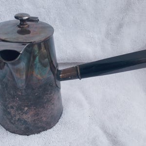 Può includere: Una caffettiera d'argento con un manico nero. La caffettiera ha una superficie testurizzata e un beccuccio laterale.