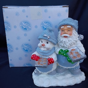 Może przedstawiać: Ceramiczna figurka bożonarodzeniowa przedstawiająca bałwana i Świętego Mikołaja trzymających tace z ciasteczkami. Bałwanek ma niebieską czapkę i szalik z czerwonymi rękawicami. Święty Mikołaj ma białą brodę, niebieską czapkę i zielone rękawice. Na pudełku widnieje napis "Snow Buddies".