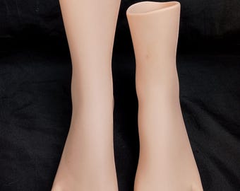 May include: Two plastic mannequin feet, one larger than the other, are shown against a black background. The feet are a pale pink color and have five toes each.