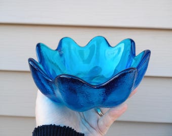 May include: A vibrant, turquoise-blue glass bowl with a unique, scalloped edge. The bowl's design resembles a flower, with each petal-like curve adding to its visual appeal. The bowl is held against a neutral background.