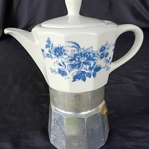 May include: A stovetop espresso maker featuring a white ceramic upper chamber decorated with a blue floral pattern and a matching lid. The lower base is constructed from metallic silver aluminium with a faceted, octagonal shape and a small valve.