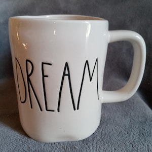 Puede incluir: Taza de cerámica blanca con asa curva. La palabra "DREAM" está impresa en negro, con letras manuscritas, en la parte delantera de la taza. La taza está sobre una superficie gris.