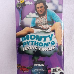 May include: A purple and white VHS tape case featuring a man in a blue shirt and floral apron. The case reads "Monty Python's Flying Circus" and "Contains: Dead Parrot, the Lumberjack Song & Albatross".