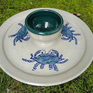 May include: A ceramic serving platter with a speckled off-white finish featuring three hand-painted blue crabs. A matching small bowl with a deep green interior is centered on the platter, creating a two-piece set for serving food.