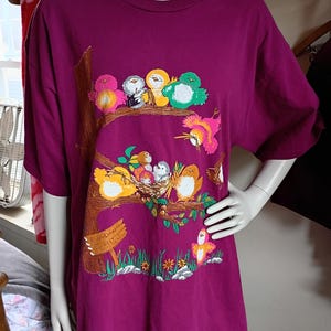 May include: A plum-colored t-shirt featuring a colorful bird design. The graphic includes a tree branch with several birds, a nest with baby birds, and a sign that reads "Building School". The shirt has short sleeves and a crew neck.