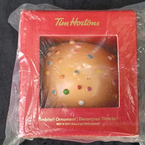 May include: A Tim Hortons Timbits ornament inside a red square box with a clear plastic window. The ornament is a round, tan-coloured pastry shape decorated with small, colourful sprinkles. The box features the text Tim Hortons and Timbits Ornament.
