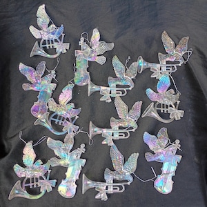 Puede incluir: Colección de adornos iridiscentes con una paloma con las alas extendidas, combinada con varios instrumentos musicales. Los instrumentos incluyen trompetas, trompas y violines. Cada adorno tiene una cuerda para colgar.