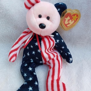 May include: A patriotic Ty Beanie Baby bear plush toy. The bear features a pink face, red and white striped ears and arms, and a body split between a blue field of white stars and red and white stripes. A gold heart-shaped tag is attached.