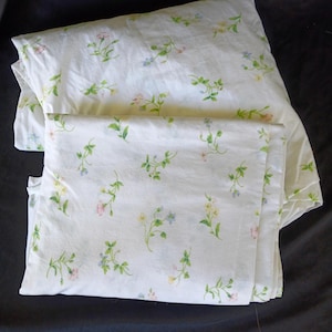 May include: A white pillowcase featuring a repeating pattern of small, delicate flowers in shades of pink, yellow, and blue, accompanied by green stems and leaves. The fabric is folded, showing the floral print across the surface.