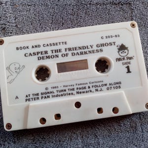 以下が含まれることがあります： 「キャスパー・ザ・フレンドリー・ゴースト・デーモン・オブ・ダークネス」とピーターパンのロゴが入った白いカセットテープ。テープには「本とカセット」と「サイド1」の文字も含まれています。