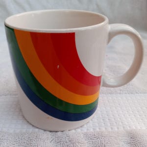 May include: White ceramic mug with a curved handle, featuring a vibrant rainbow design on the side. The rainbow arcs from red to orange, yellow, green, and blue. A simple, everyday coffee or tea mug.