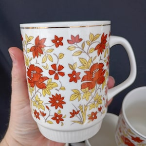 Può includere: Tazza in ceramica bianca con un motivo floreale rosso, arancione e giallo. La tazza ha un bordo dorato e un manico. Sono visibili altre tazze con motivi simili. Le tazze sono tenute da una persona.