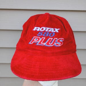 May include: A red corduroy hat with the text "ROTAX 580 PLUS" embroidered in white and blue. The hat has a rounded crown and a wide brim. The hat is held up by a hand.