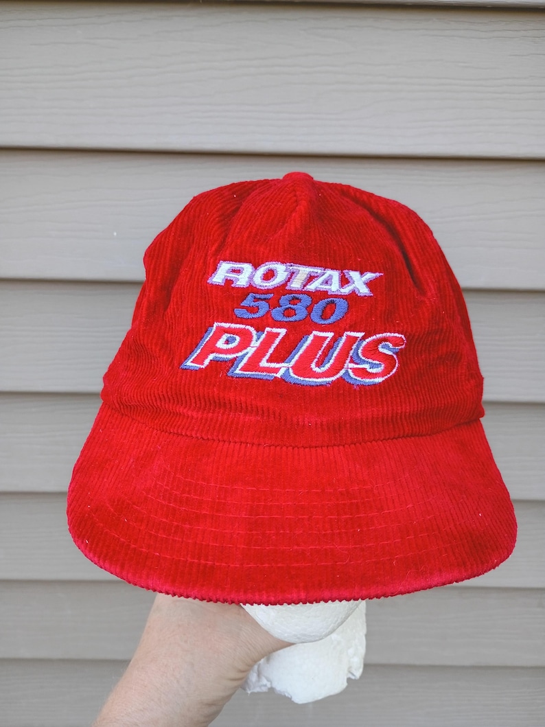 May include: A red corduroy hat with the text "ROTAX 580 PLUS" embroidered in white and blue. The hat has a rounded crown and a wide brim. The hat is held up by a hand.