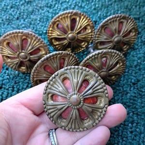 Peut inclure: Un ensemble de six boutons de placard ronds dotés d'un cadre en métal doré avec un motif radial décoratif et une incrustation rouge orangé visible à travers les découpes. Chaque bouton présente un bord texturé et un détail central circulaire.