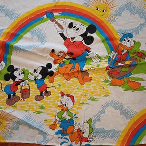 May include: A white fabric featuring a colorful cartoon print of Mickey Mouse, Pluto, Donald Duck, Daisy Duck, and three ducklings painting a rainbow. The scene includes a yellow sun, blue clouds, and a yellow cobblestone path on a white background.
