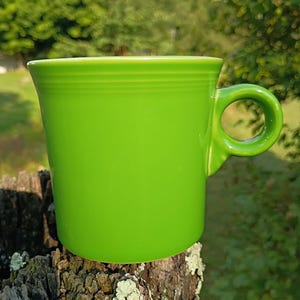 May include: A bright lime green ceramic mug featuring a smooth, cylindrical body and a circular handle on the right side. The mug has a series of subtle, horizontal embossed rings circling the upper portion of the exterior wall, providing a simple texture to the otherwise solid-coloured, glossy finish of the drinkware item.