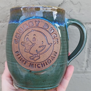 May include: Green ceramic mug with a handle, featuring a brown circular emblem with the words "CHUBBY DUCK" and "FLINT MICHIGAN" surrounding a cartoon duck. The rim has a blue and brown glaze.