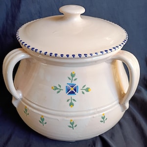 May include: Cream-colored ceramic casserole dish with a lid. The dish features two handles and is decorated with blue, yellow, and green floral patterns. The lid has a blue dotted border.