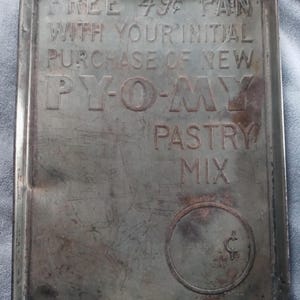 Op de afbeelding: Een vintage metalen bakvorm met de tekst "FREE 49¢ PAN WITH YOUR INITIAL PURCHASE OF NEW PY-O-MY PASTRY MIX" in reliëf op de voorkant. De vorm heeft een cirkelvormig logo met een centsymbool in het midden.