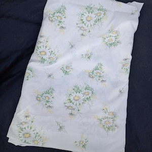 Puede incluir: Tela blanca con un estampado floral de margaritas. Las margaritas tienen centros amarillos y hojas verdes. La tela está doblada y parece una sábana u otra prenda de cama.