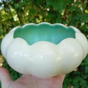 May include: A round ceramic bowl with a scalloped edge, featuring a glossy cream-colored exterior and a smooth, vibrant turquoise interior. The bowl has a wide, open top and a curved, rounded body, showcasing a two-tone color scheme.