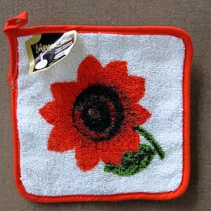 May include: A square white terry cloth pot holder with a bright orange sunflower graphic featuring a dark center and a green leaf. The pot holder has a vibrant orange fabric border and a small hanging loop. A Morgan Jones brand tag is attached.