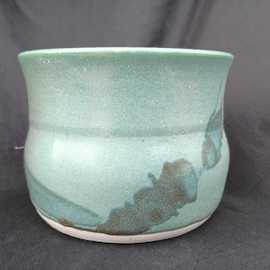 May include: A handmade ceramic mug with a teal glaze and brown accents. The mug has a rounded shape with a slightly flared rim and a white base. The mug is suitable for coffee, tea, or other beverages.