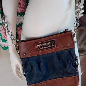 May include: A Chaps crossbody bag in brown and navy blue, with a silver chain strap. The bag features a brown top and bottom, with a navy blue center section. The Chaps logo is displayed on a rectangular plaque.