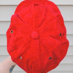 May include: A bright red corduroy baseball cap. The cap has a rounded crown with a button on top and a curved brim. It features six ventilation eyelets with metal grommets.