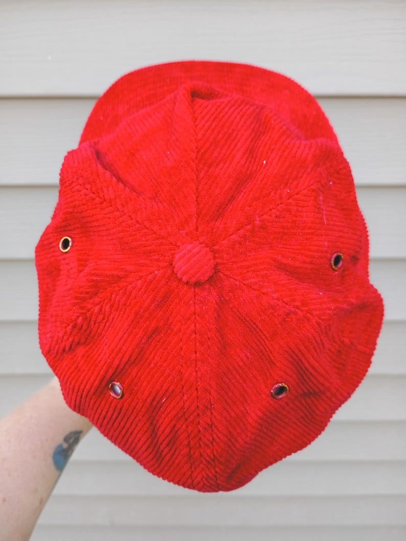May include: A bright red corduroy baseball cap. The cap has a rounded crown with a button on top and a curved brim. It features six ventilation eyelets with metal grommets.
