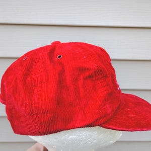 May include: A bright red corduroy baseball cap with a curved brim. The cap has a structured crown with ventilation eyelets. The hat is displayed against a light-colored background.