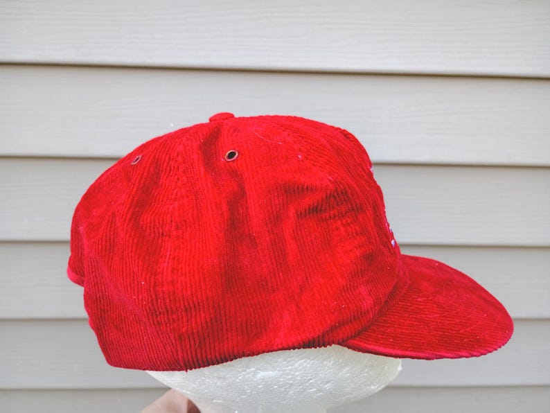May include: A bright red corduroy baseball cap with a curved brim. The cap has a structured crown with ventilation eyelets. The hat is displayed against a light-colored background.