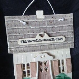 Puede incluir: Una placa de cerámica con forma de casa con fondo blanco y bordes marrones. El techo es gris con un patrón de ladrillo marrón. La placa tiene un letrero que dice "This Home Is Protected By PMS".