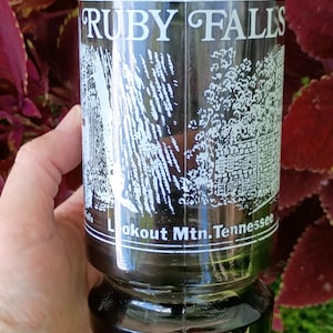 May include: A clear glass tumbler featuring a white printed graphic of Ruby Falls and the text "RUBY FALLS" at the top and "Lookout Mtn. Tennessee" at the bottom. The glass has a thick, weighted base and a cylindrical shape with a vertical seam.