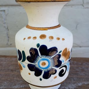 Puede incluir: Un jarrón de cerámica blanco con borde y base acampanados. El jarrón presenta un diseño floral pintado a mano en tonos azules, marrones y verdes. Puntos marrones rodean el jarrón cerca de la parte superior. El jarrón mide unos 15 cm de alto.