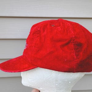 May include: A bright red corduroy baseball cap with a curved brim. The cap has a structured crown and a small embroidered design on the side. The hat is displayed on a white mannequin head against a light gray background.