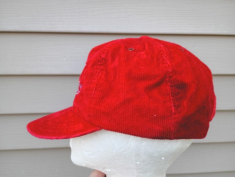 May include: A bright red corduroy baseball cap with a curved brim. The cap has a structured crown and a small embroidered design on the side. The hat is displayed on a white mannequin head against a light gray background.