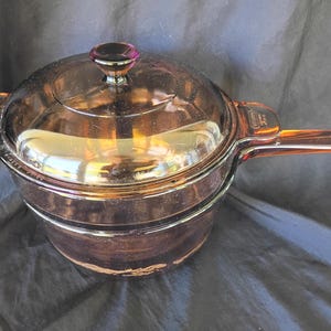May include: A translucent amber-coloured glass saucepan with a matching lid. The cookware features a long handle and a smaller side handle, both integrated into the glass body. The word Vision is embossed on the handle, and the lid has a knob.