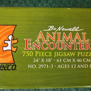 May include: A green rectangular jigsaw puzzle box featuring the text Bo Newell ANIMAL ENCOUNTERS 750 Piece Jigsaw Puzzle. The box displays the dimensions 24 inches by 18 inches, the product number 2971-3, and the age recommendation of 12 and up.