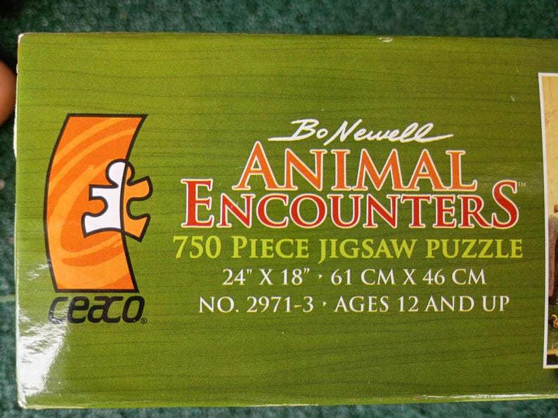 May include: A green rectangular jigsaw puzzle box featuring the text Bo Newell ANIMAL ENCOUNTERS 750 Piece Jigsaw Puzzle. The box displays the dimensions 24 inches by 18 inches, the product number 2971-3, and the age recommendation of 12 and up.