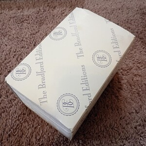 May include: A rectangular box with a light beige surface. The box is printed with the words "The Bradford Editions" and a circular emblem in a repeating pattern. The box is sealed with clear tape.