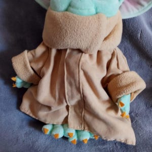 May include: A soft toy of a character from a science fiction franchise. The toy features a light brown robe, mint green skin, and large ears. The character's hands and feet are mint green with orange claws. The toy is lying on a blue surface.