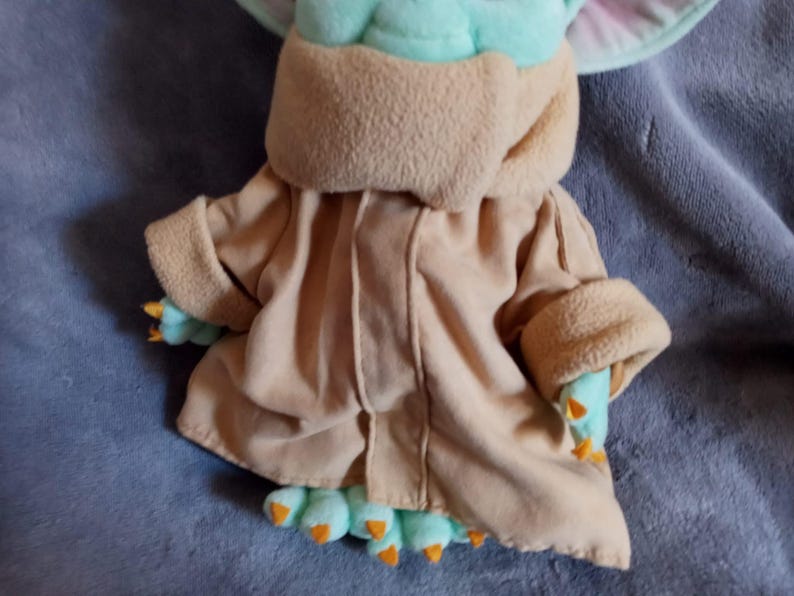 May include: A soft toy of a character from a science fiction franchise. The toy features a light brown robe, mint green skin, and large ears. The character's hands and feet are mint green with orange claws. The toy is lying on a blue surface.