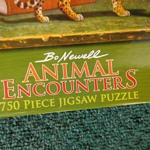 May include: A green cardboard box for a 750 piece jigsaw puzzle titled Bo Newell Animal Encounters. The box features white and orange text on a green background, with a partial view of a leopard and a small brown creature visible at the top edge.