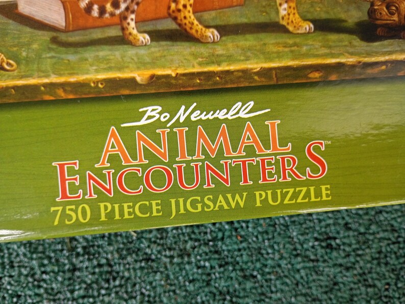 May include: A green cardboard box for a 750 piece jigsaw puzzle titled Bo Newell Animal Encounters. The box features white and orange text on a green background, with a partial view of a leopard and a small brown creature visible at the top edge.