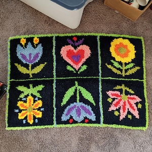 Puede incluir: Una alfombra rectangular negra con un borde verde, con seis diseños florales bordados. Las flores son de varios colores, incluyendo azul, amarillo, naranja y rosa. La alfombra está colocada sobre una alfombra marrón.