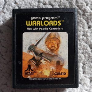 May include: A black Atari game cartridge with a picture of a knight on horseback. The game is called "Warlords" and is a game program. The cartridge is labeled "CX2610" and has the Atari logo. The text on the cartridge reads "Use with Paddle Controllers".